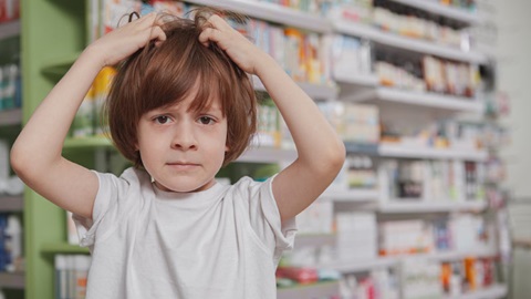 Menino de cabelo comprido ruivo, com as mãos na cabeça e ar preocupado, em frente a uma estante com medicamentos, numa farmácia